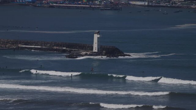 Wide view of Los Yuyos beach with lighthouse on a summer day, people enjoying sea and shore in slow motion, distant figures without visible faces along the Lima Peru coast