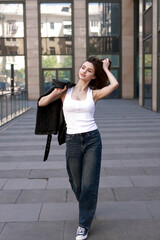 Fototapeta premium A beautiful brunette in a leather jacket, jeans and sneakers walks down the street of a modern city.