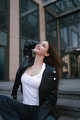 Fototapeta premium A beautiful girl sits on the steps of a modern building in a leather jacket and jeans.
