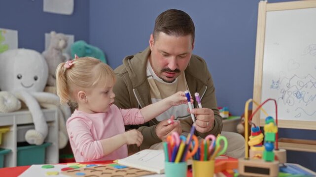 Man guiding young girl to hold markers and color on paper at a preschool building while seated at a craft table strewn with toys and pencils; learning patience.