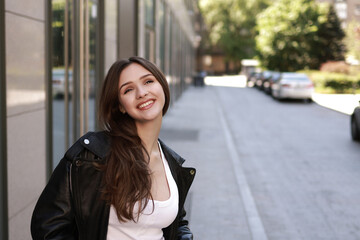 Fototapeta premium A beautiful brunette girl in a leather jacket, jeans and sneakers on the street of a modern city.