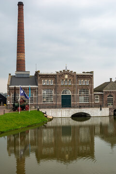 View of the Dutch Steam Engine Museum with its tall brick chimney and historic facade reflected in the canal water in Medemblik, Noord-Holland, Netherlands.