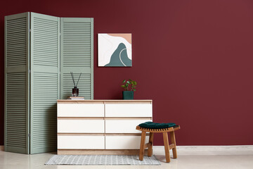 Obraz premium Interior of living room with folding screen, chair and home decor on commode near burgundy wall