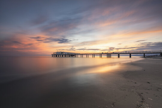 View of the modern pier extending into the calm sea under a vibrant sunset sky with orange and purple clouds over the sandy beach in Lido di Camaiore, Tuscany, Italy.