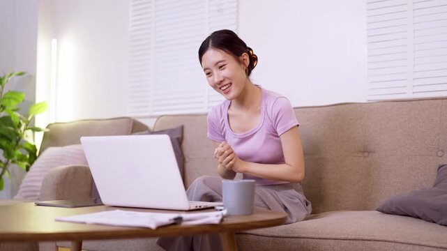 young Asian women remote work from home using laptop computer video call online meeting sitting sofa living room freelance digital nomad business casual