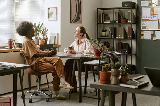 African American young adult woman and Caucasian young adult woman meeting at office desk, discussing project over coffee. Ideal for teamwork, mentoring, workplace communication