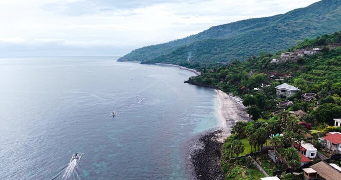 Drone view of the peaceful coastline in Amed, Bali, featuring calm ocean waters, beachfront villas, fishing boats, and lush green hills along the tropical shoreline of Indonesia.