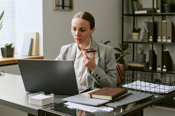 Fototapeta premium Caucasian middle aged businesswoman speaking into smartphone while working on laptop at desk, managing remote communication. Ideal for office workflow, client support, hybrid work