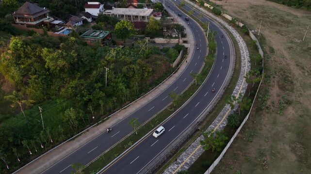 Bird's eye drone view of a winding multi lane road with cars and scooters moving through lush countryside, elegant homes and trees glowing in warm sunset light, scenic travel scene