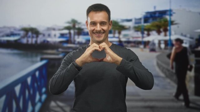 Man forms a heart with hands on a seaside street, smiling and winking while wearing a dark sweater and wristwatch; friendly affection.
