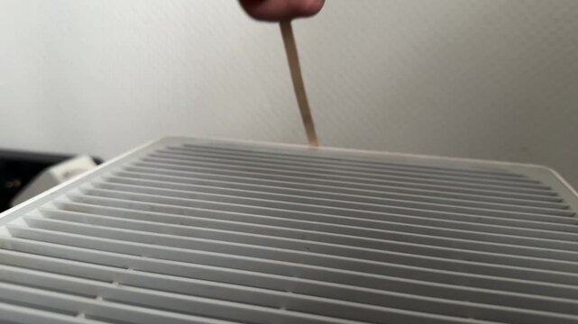 Person sliding wooden stick along air purifier grille close up at home, producing vibrating sound, improvised interaction, everyday object experiment concept
