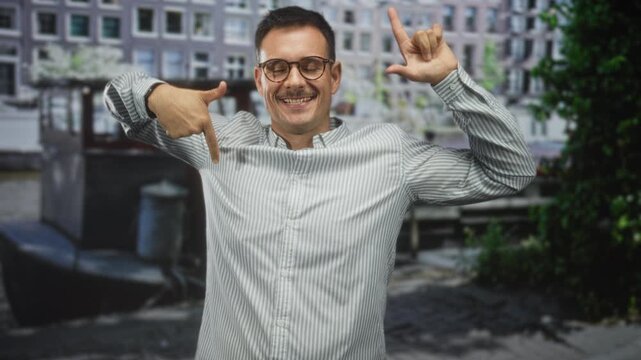 Man with moustache and glasses points finger down and forms l with other hand on street; playful confidence.