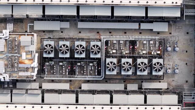 Aerial view of industrial cooling units on rooftop. Large fans rotate steadily in rows. Metal structures dominate the concrete surface