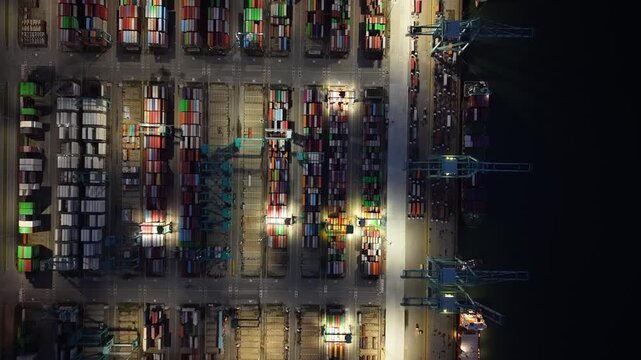 Night aerial drone view of busy container terminal with illuminated cranes, stacked containers, and operations.