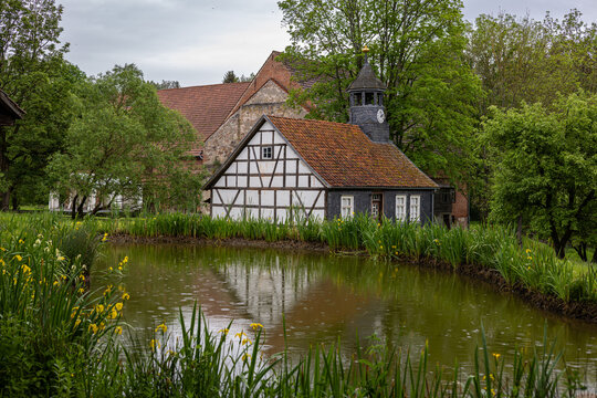 View of the Hennebergisches Museum Kloster Vessra, featuring a half-timbered house with a clock tower reflected in a pond with yellow irises Kloster Vessra, Thuringen, Germany.