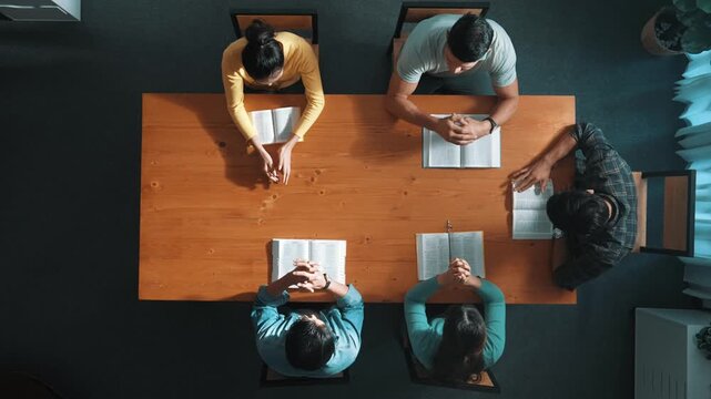 Top down aerial view of smart team hold hands on the holy bible and praying god while sitting at meeting table. Group of people hand folded in prayer with faith, spirituality and religion. Symposium.
