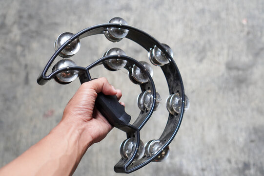 A close-up shot of a hand holding a black, crescent-shaped plastic tambourine against a blurred, neutral grey concrete background.