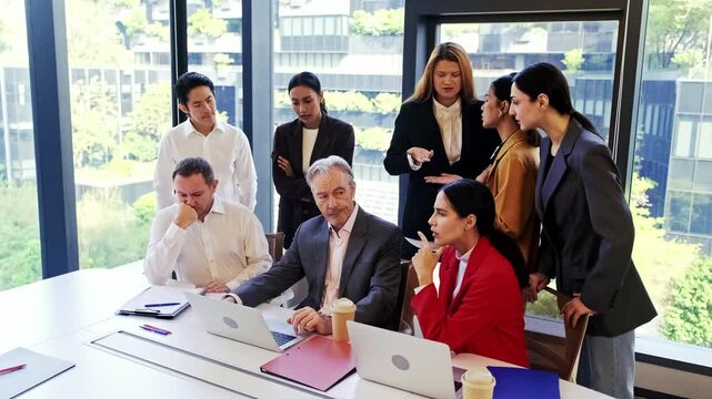 Team of professionals gathered around laptops in a modern office, reviewing data and discussing strategy by the window during a focused meeting.