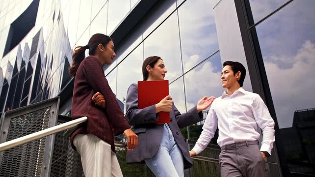 Three professionals converse outside a modern glass office building, standing on steps, holding documents and smiling during a casual meeting.