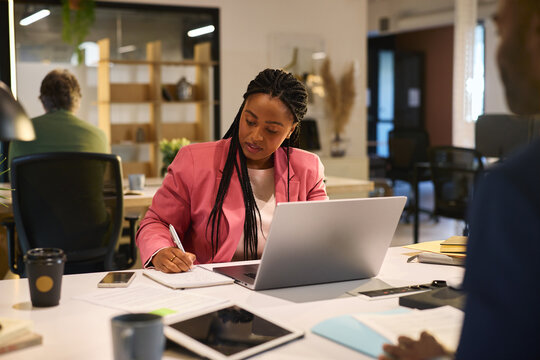 Serious black woman writing notes while working on laptop at creative coworker space