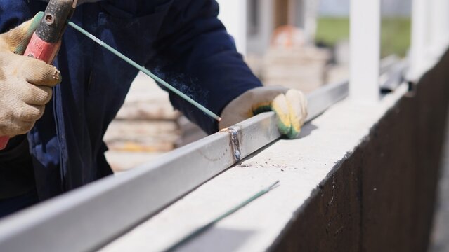 Closeup Metalworker Welding Joint, Sparks And Molten Bead Visible, Gloved Hand Steadying Bracket On Metal Profile, Industrial Site Background, Precise Joiner Action And Heat Intensity
