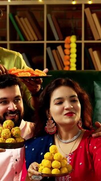 Friends wishing Happy Diwali to camera, Indian young couple friends in traditional festive wear celebrating Hindu festival at decorated home, holding laddu barfi while smiling greeting joyfully