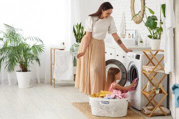 Young woman and her daughter setting up washing machine with dirty clothes in laundry room