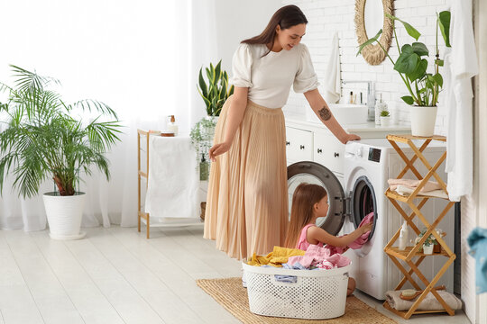 Young woman and her daughter setting up washing machine with dirty clothes in laundry room