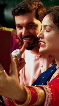 Taking Diwali selfie with friends, Indian Asian young married couple in traditional attire celebrating Hindu festival Deepavali at decorated home holding laddu barfi sweets clicking selfie