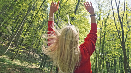 A caucasian woman stands in a vibrant forest, arms raised towards the sun filtering through the lush green canopy, her long blonde hair swaying in the breeze. © wkproduction