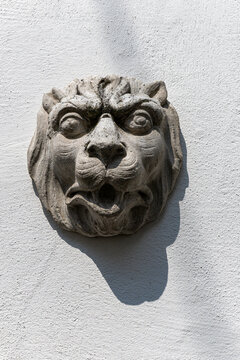 View of a stone lion head sculpture with an open mouth mounted on a white textured wall under bright sunlight Monreal, Rhineland-Palatinate, Germany.