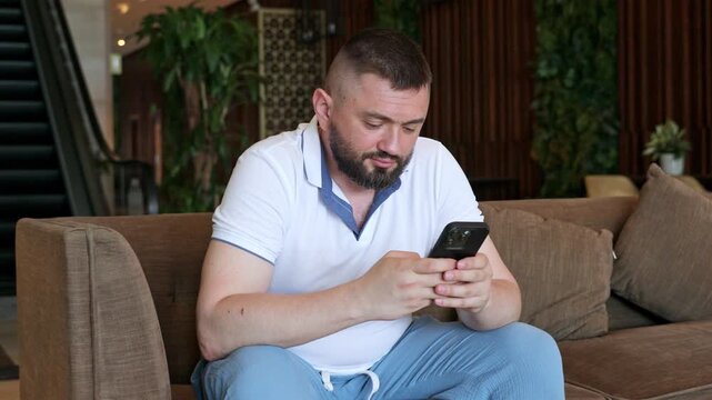 Side view young man with a beard dressed in a white polo shirt and light blue pants sits on a couch, focused on his smartphone in a contemporary indoor space featuring escalators and plants