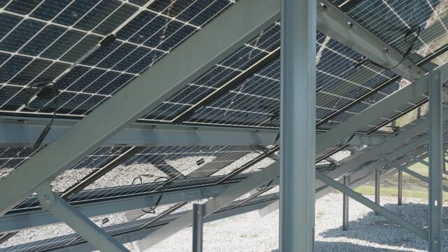 Industrial steel mounting structures and racks support bifacial solar panels at farm. Rear view of dual sided photovoltaic modules for renewable energy production on white gravel ground.