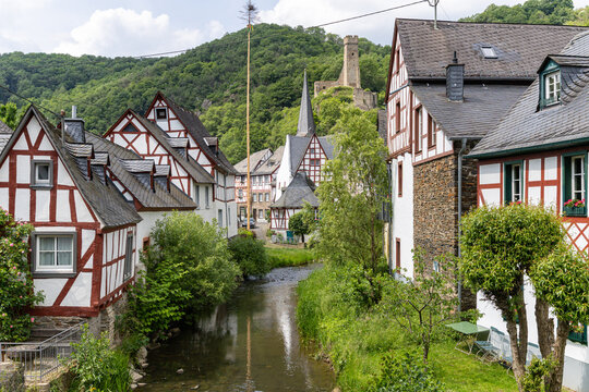 View of historic half-timbered houses along a narrow river with a castle ruin on a green hill under a bright sky Monreal, Rhineland-Palatinate, Germany.