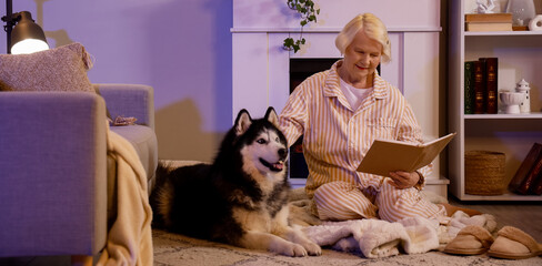 Senior woman with Husky dog reading book near fireplace at home late in evening © Pixel-Shot