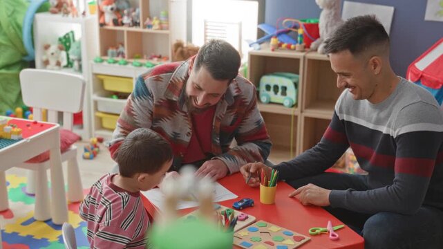 Boy drawing with pencil at a red activity table while two men guide and point in a daycare building; parental support joyful.
