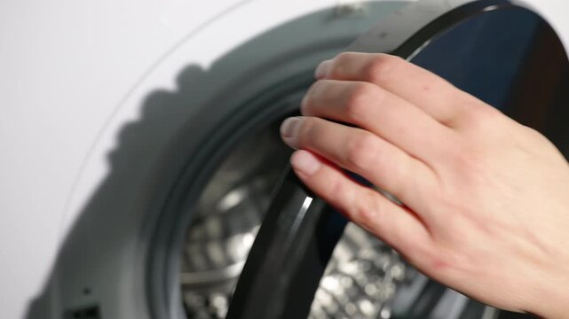 Woman opening washing machine door revealing empty drum. Close-up view of a woman's hand opening the door of a modern washing machine, revealing the empty, clean, and shiny stainless steel drum