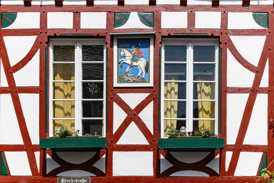View of a traditional half-timbered house facade featuring a Saint George icon, red timber framing, and green window boxes in Monreal, Rhineland-Palatinate, Germany.