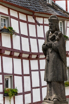 View of a stone statue of a religious figure in front of a traditional half-timbered house with white walls and dark wooden beams Monreal, Rhineland-Palatinate, Germany.