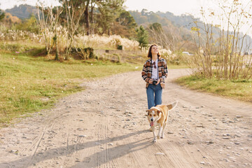 Fototapeta premium A woman and her dog walk along a dirt path in a sunny countryside setting, wearing a plaid shirt and jeans, as they move through an open field and trees in a peaceful outdoor moment.