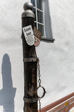 View of the historic Schandbaum pillory with iron shackles and a coat of arms against a white building wall in Monreal, Rhineland-Palatinate, Germany.