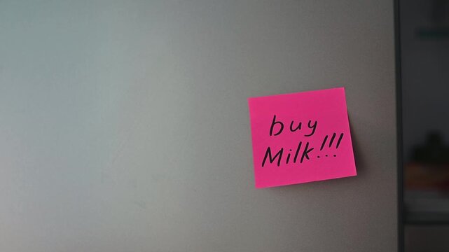 A hand places a bright pink sticky note on a refrigerator door. The note has a shopping reminder written on it, indicating what needs to be purchased