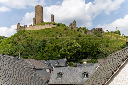 View of the historic Monreal Castle ruins on a lush green hill above traditional slate rooftops under a cloudy sky in Monreal, Rheinland-Pfalz, Germany.