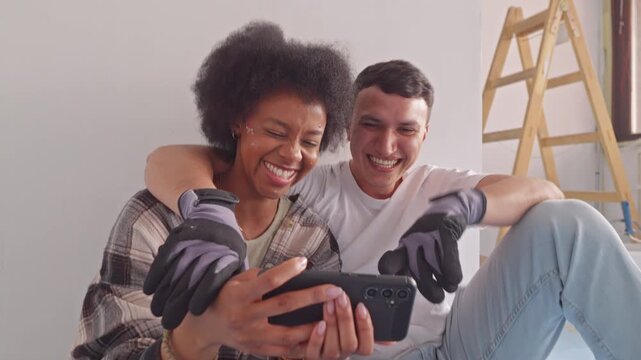 A smiling couple, surrounded by laughter, shares a moment over a smartphone. The man and woman, both engaged in housework, wear gloves, hinting at recent or ongoing renovation.