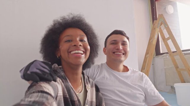 A young couple smiles amidst their home renovation, paint splatters lightly on the woman's face as she wears gloves and stands close to her partner, symbolizing their fun and effort.