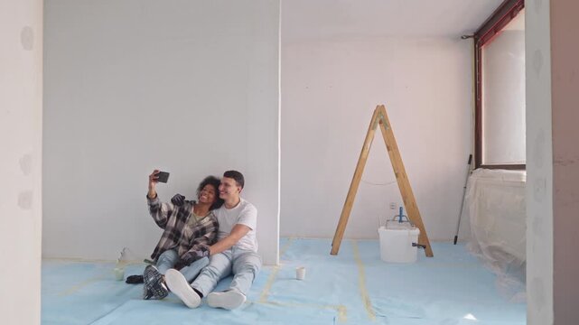 In a sunlit room under renovation, a young couple captures their moment, legs intertwined on a protective blue sheet, with freshly plastered walls and DIY supplies nearby.