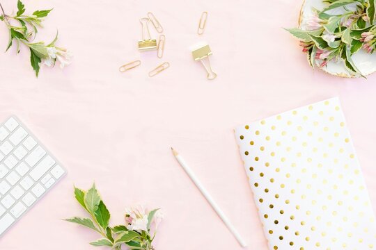 Flat lay of feminine desk with keyboard notebook and floral elements minimalist workspace for creative professionals and remote workers with pastel pink background and gold accents