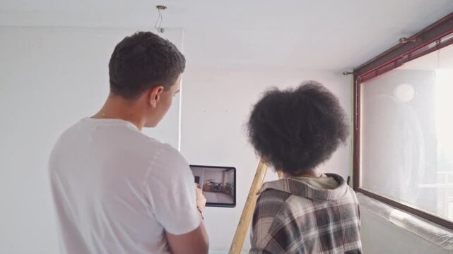 A couple in casual attire examines a tablet in a room with unfinished walls, collaborating on a home renovation project, showcasing their teamwork and shared vision for the interior.