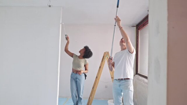 A couple diligently paints a room's ceiling during their home renovation project. The woman focuses on a spot on the wall, while the man, on a small step ladder, stretches to paint higher areas.