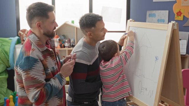 Two men and little boy drawing on a whiteboard easel, one man guiding marker while the other watches in a playroom building; playful bonding.